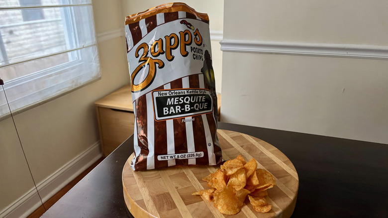 Bag of Zapp's Mesquite Bar-B-Que chips on cutting board with handful of chips