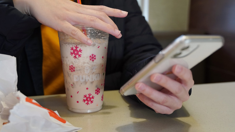 Young woman scrolls on her iPhone 11 as she sips on an iced drink from Dunkin Donuts