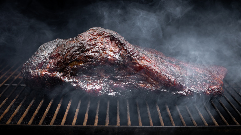 Smoke rising around a slow cooked beef brisket on the grill grates of a smoker barbecue