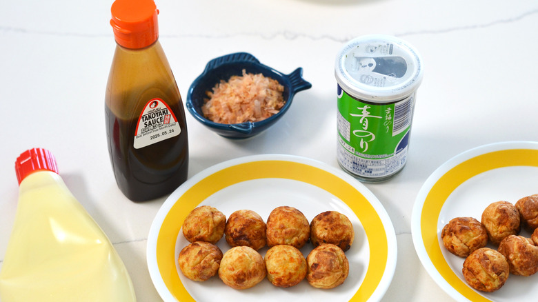 Plate of cooked takoyaki surrounded by toppings: takoyaki sauce, mayo, aonori, katsuobushi