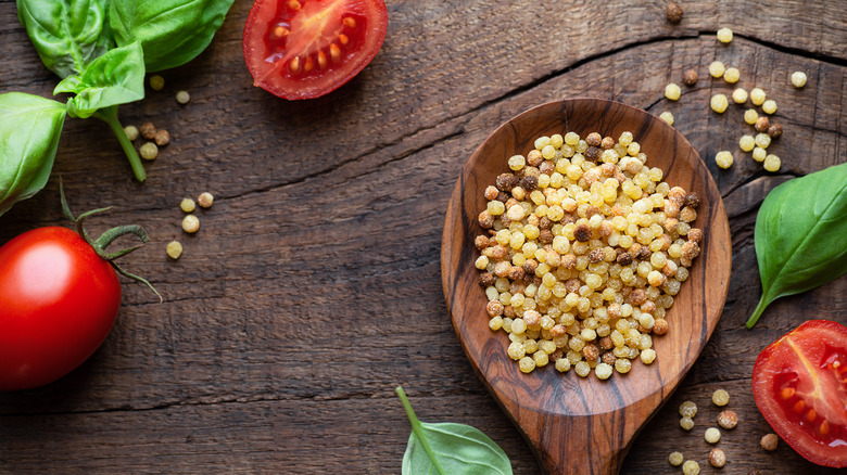 beads of fregola in a wooden spoon and strewn about a wooden table with halves of cherry tomatoes and leaves of basil