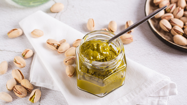 Jar of pistachio butter with spoon surrounded by whole pistachios