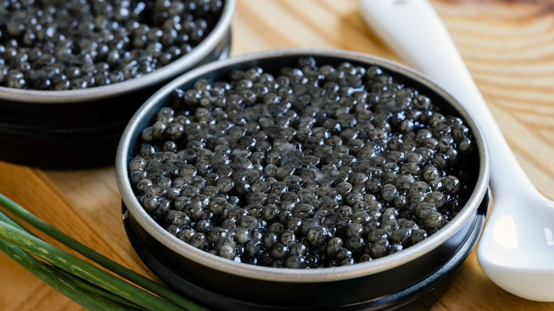 Two open tins of caviar sit on a wooden board with a white spoon and green onions on the side