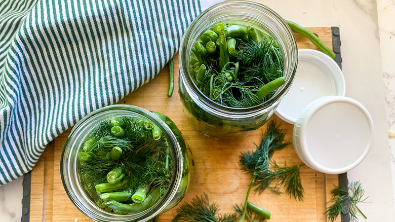 two mason jars with green beans and pickling spices on a wood cutting board