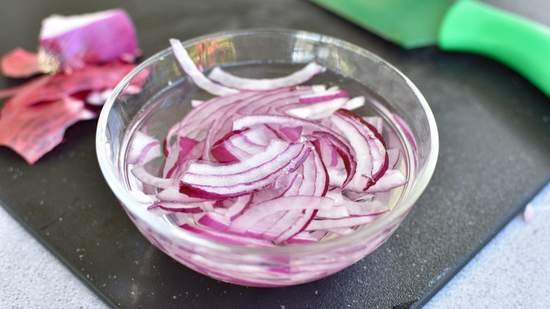 A glass bowl filled with water and sliced red onions next to a green knife