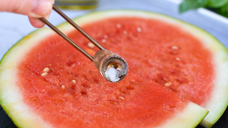 A hand holding a metal measuring spoon of salt over slices of watermelon