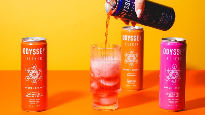 Person pouring a can of Odyssey Elixir into a glass cup surrounded by other Odyssey canned drinks