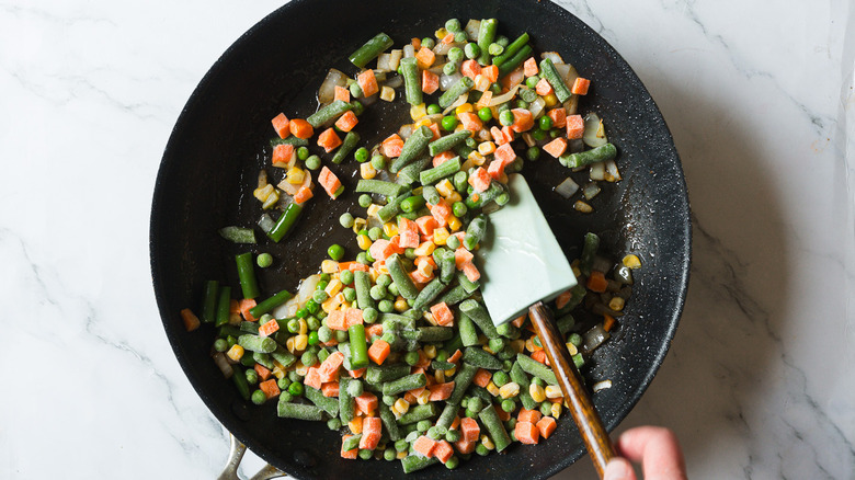 Hand mixing frozen veggies in pan with spatula