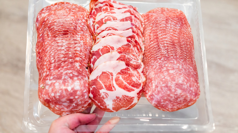 A hand holding a plastic tray filled with slices of deli meats