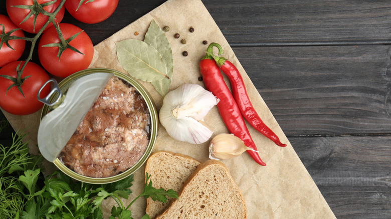 canned meat with the lid peeled back and chili peppers, fresh herbs, tomatoes, and garlic around it