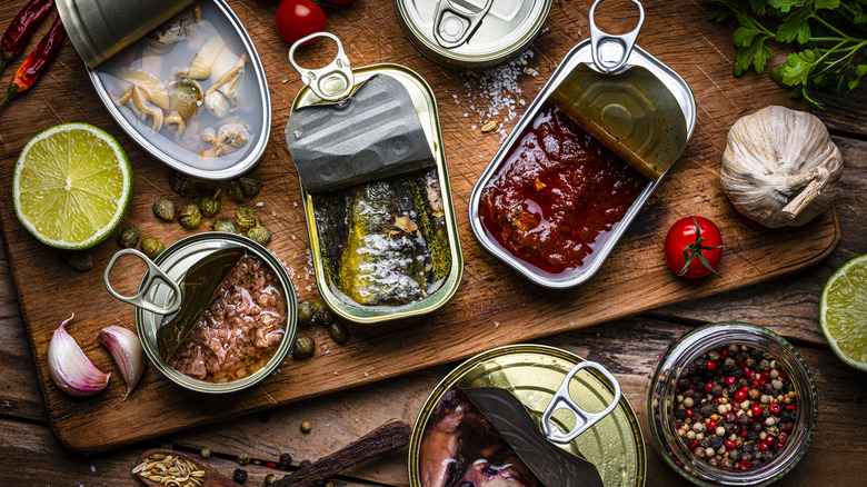 various open tins of meat on a wood board with fresh ingredients and spices sprinkled around