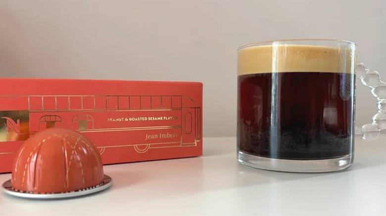 A sleeve of Nespresso Barista Creations Peanut & Roasted Sesame coffee pods with a pod in front of it pictured next to a glass mug of coffee