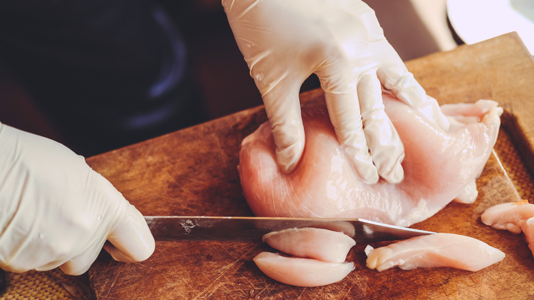 Gloved hands slicing raw chicken on cutting board