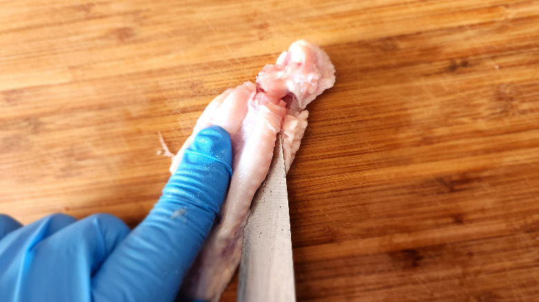 gloved hand holding a chicken wing on a bamboo cutting board, as a knife slices it lengthwise along the bone