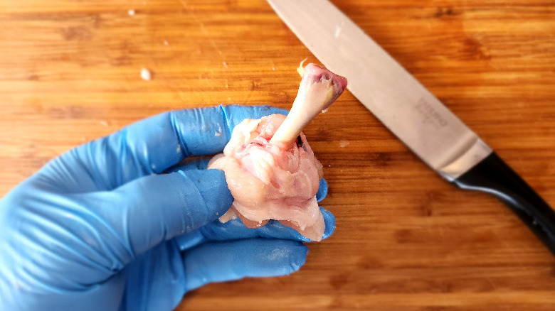 gloved hand holding a "frenched" chicken wing drumette over a bamboo cutting board, with boning knife in the background