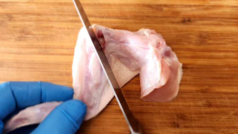 gloved hand holding a chicken wing on a bamboo cutting board, as a knife severs the drumette from the wing