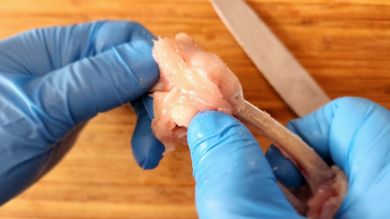 gloved hands baring the bones of a chicken wing, knife and cutting board in background