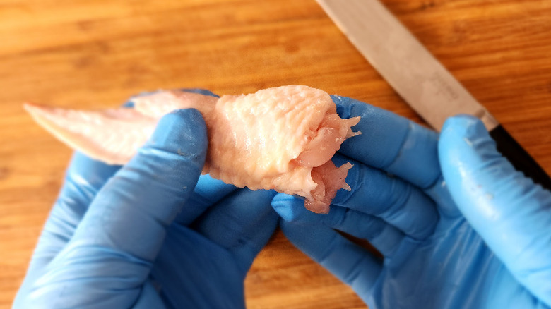 gloved hands holding a newly deboned chicken wing, knife and cutting board in background