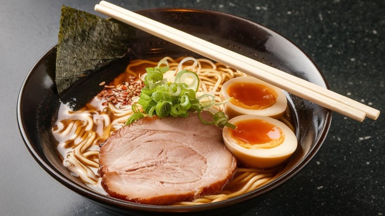 tonkotsu ramen dish with jammy egg, seaweed sheet, scallions, red pepper flakes, pork and noodles, in a black bowl with chopsticks