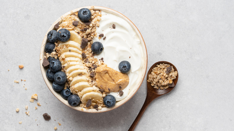 A bowl of yogurt with banana, blueberries, yogurt, and peanut butter next to a spoon of granola