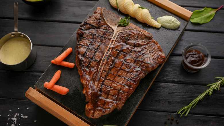 Porterhouse steak resting on cutting board surrounded by veggies, greens, and sauces