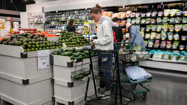 A woman is wearing headphones while looking at avocados in the grocery store.