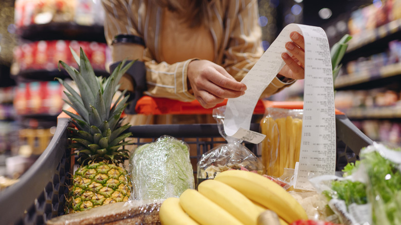 A woman standing behind a grocery cart of food reviews a long grocery receipt.