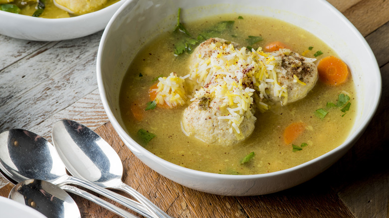 Matzo ball soup garnished with herbs, cracked black pepper, and cheese in a white bowl