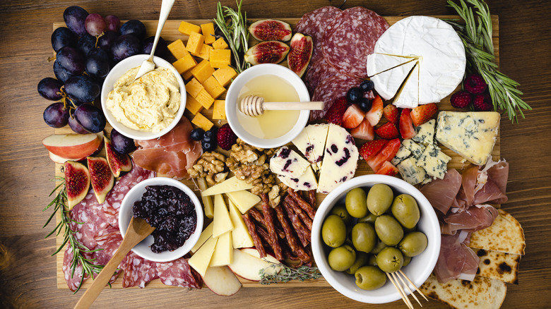 Top-down view of charcuterie board with grapes, cheeses, meats, nuts, dips, olives, and honey spread out across a wooden board