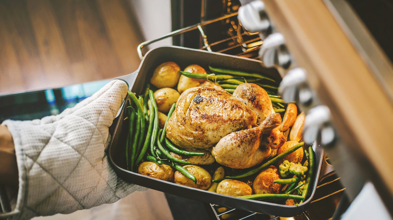 Hands in oven mitts taking roast chicken, potatoes, and green beans out of oven