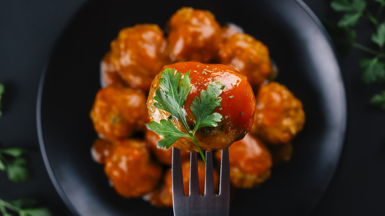 Saucy meatball on fork behind plate of meatballs