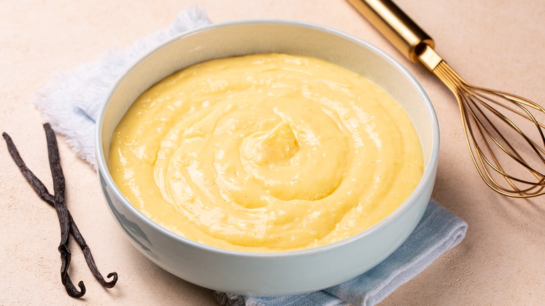 Pastry cream in a bowl surrounded by vanilla beans and a whisk