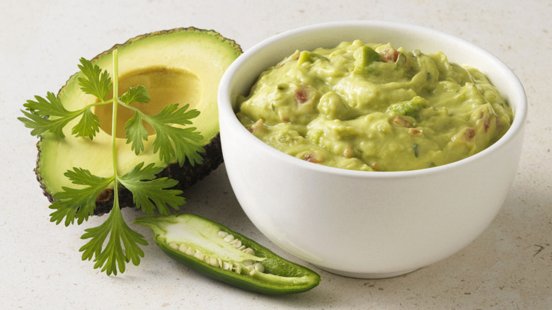 Guacamole in a white bowl beside halved avocado and herb sprig