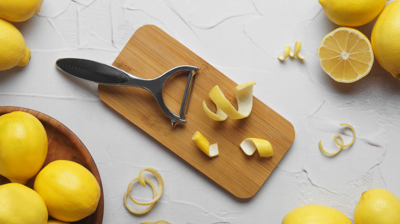 A vegetable peeler on a wooden chopping board surrounded by lemon peels and whole lemons