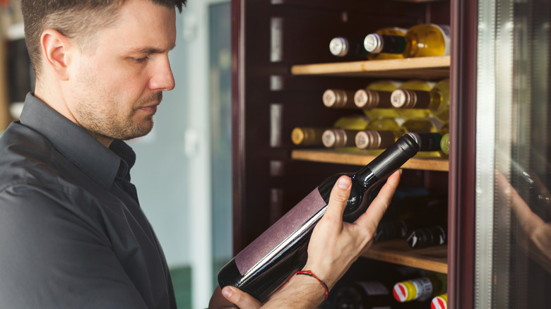 man inspecting a bottle of wine in front of a wine refrigerator