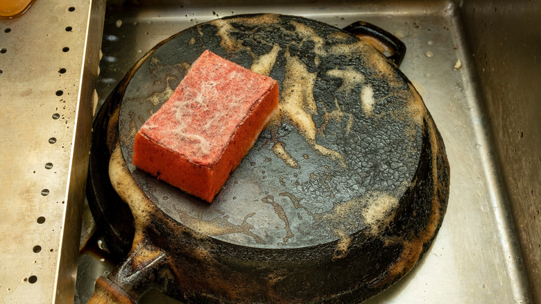 A soapy sponge rests on a dirty cast iron skillet