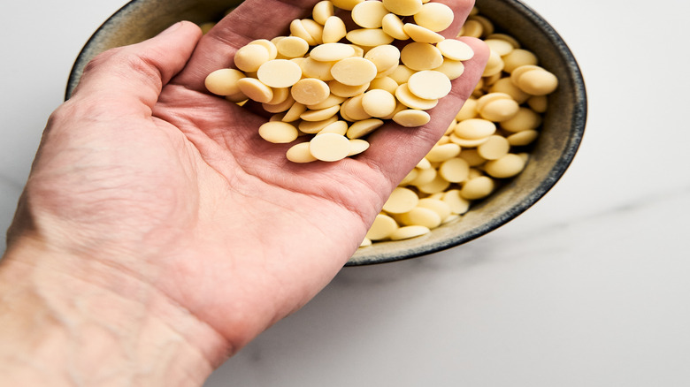A hand holds a handful of white chocolate morsels above a bowl.