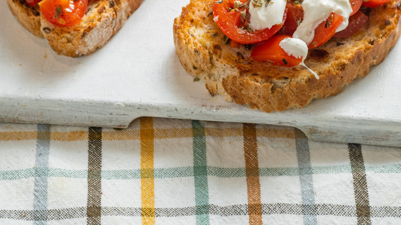 Three slices of toasted bread topped with creamy burrata, vibrant roasted cherry tomatoes, and fresh basil leaves. The crostini are arranged on a white serving board alongside a bowl of extra tomatoes and a small dish of burrata garnished with olive oil and basil, creating a fresh and colorful presentation.