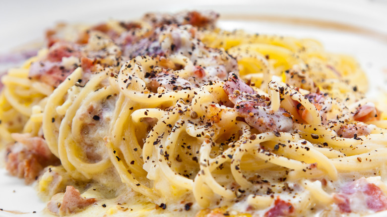 A close-up of creamy spaghetti carbonara topped with crispy guanciale and generously sprinkled with freshly cracked black pepper. The pasta glistens with its silky, egg-based sauce, while bits of golden-brown guanciale add texture and flavor. The dish is plated simply.