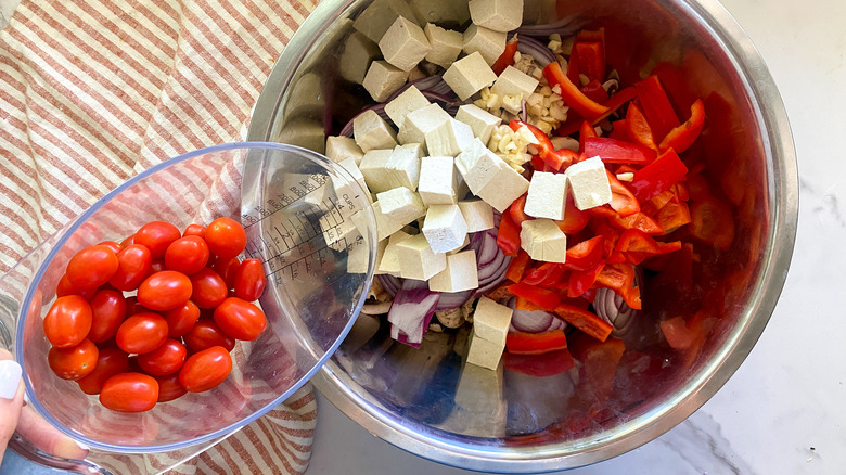 Pouring tomatoes into bowl of tofu and vegetables.