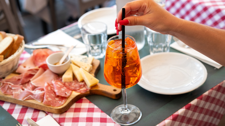 Mortadella as part of an Italian salumi spread, served with an Aperol spritz with straw grabbed by woman's hand