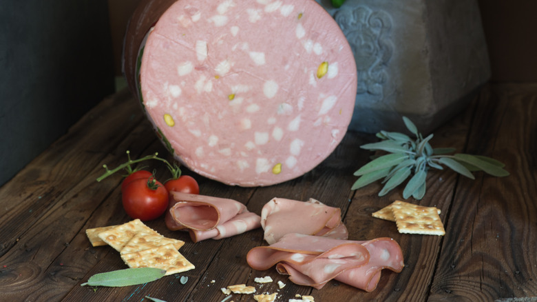 Mortadella in log form and shaved, against a wooden cutting board with crackers and tomatoes