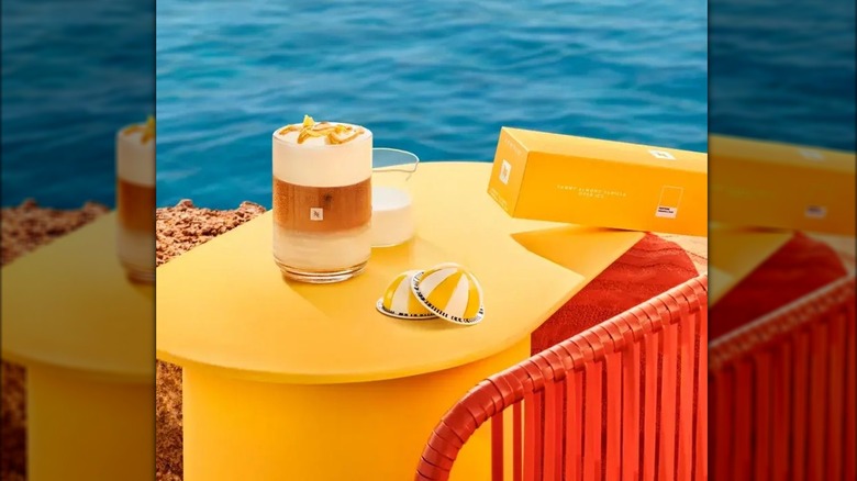 A mug of coffee next to orange and white striped Nespresso capsules on a table near an ocean