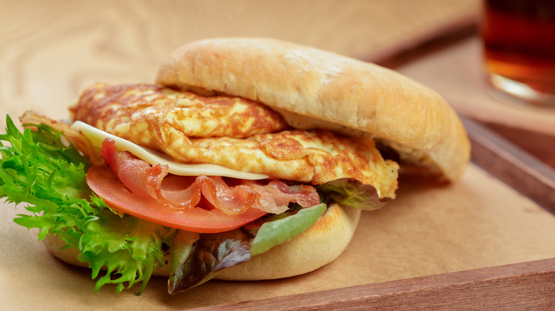 omelete sandwich with sliced tomato, lettuce, bacon, and cheese on a wood board