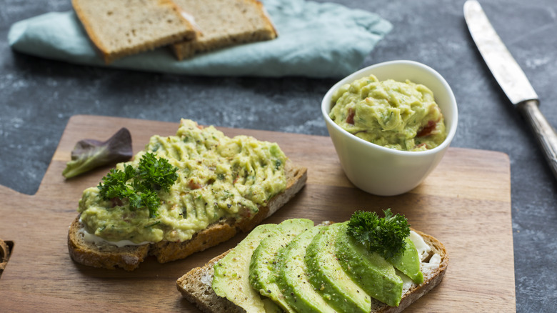 A cutting board with 2 slices of avocado toast and sliced bread in the background