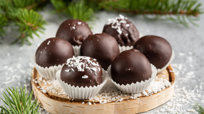 A log shaped plate of chocolate peanut butter balls dusted in coconut with ferns surrounding it