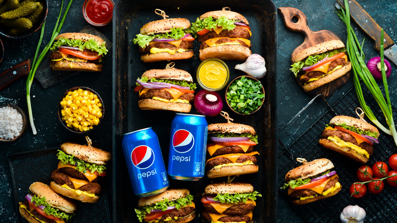 an array of burgers, pickles, and condiments with two cans of pepsi