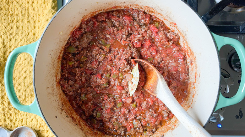 Ground beef, tomatoes, peppers, and spices in large pot on stove top with wooden spoon