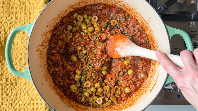 Stirring olives, raisins, and capers into Cuban picadillo in pot on stovetop with wooden spoon