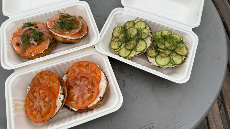 Three kinds of bagels in takeout containers, lox and cream cheese, tomato and cream cheese, cucumber and white fish spread